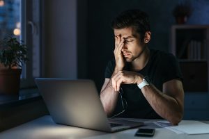 Young businessman has headache while working overtime with laptop at home office late night. Stressed depressed freelancer touching his head, feeling pain in eyes. Insomnia, internet addiction, work late
