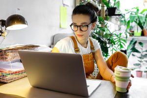 Young female gardener in glasses using laptop, communicates on internet with customer in home garden/greenhouse, hold reusable coffee/tea mug.Cozy office workplace, remote work, stay home concept
