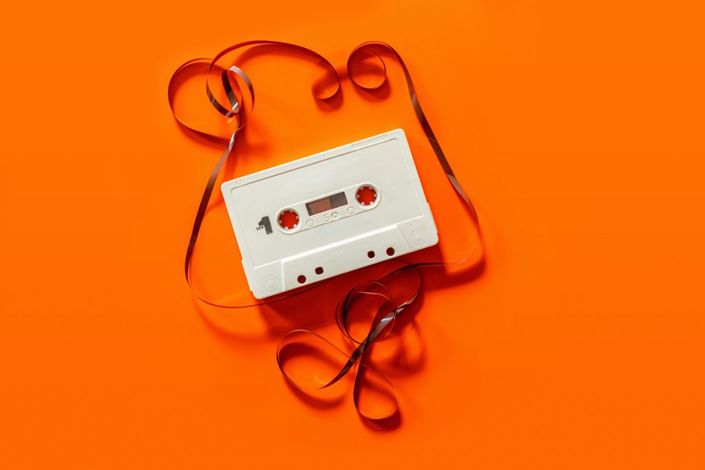 A plain white cassette tape lying flat on a bright orange surface with the tape pulled out and curling around the cassette.